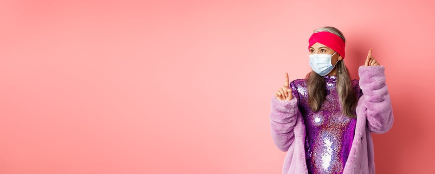 Covid-19, social distancing and fashion concept. Cheerful asian senior woman likes to party, dancing in respirator and shiny purple dress, standing over pink background photo