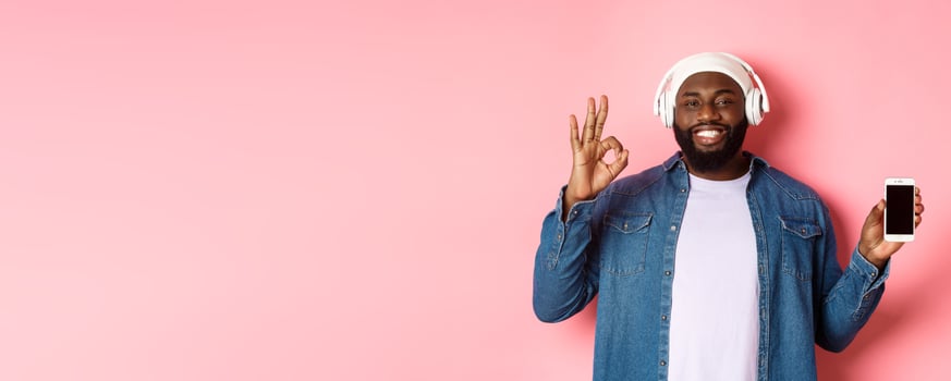 Happy hipster guy listening music on headphones and showing mobile screen, smiling satisfied, showing okay sign in approval, like playlist, standing over pink background photo