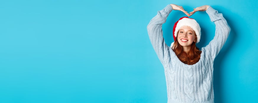 Winter holidays and Christmas Eve concept. Cute redhead teen girl in santa hat and sweater, making heart sign and smiling, wishing merry xmas, standing over blue background photo