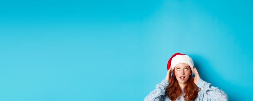 Winter holidays and Christmas eve concept. Head of cute redhead girl in santa hat, appear from bottom and looking up at copy space, staring logo, standing over blue background photo