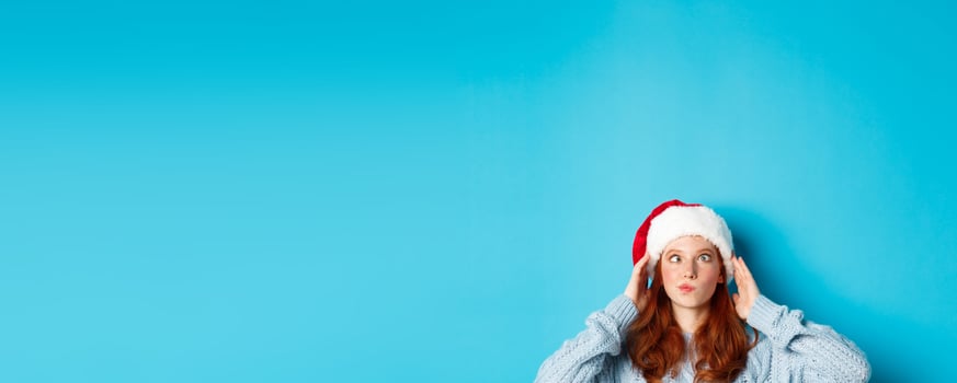 Winter holidays and Christmas eve concept. Head of funny redhead girl in santa hat, appear from bottom and squinting, making silly faces, standing near copy space on blue background photo