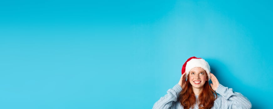 Winter holidays and Christmas eve concept. Head of cute redhead girl in santa hat, appear from bottom and looking up at copy space, staring logo, standing over blue background photo