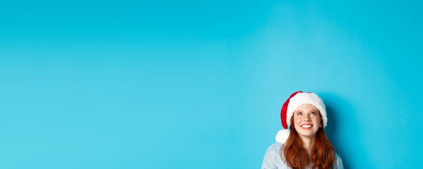 Winter holidays and Christmas eve concept. Head of cute redhead girl in santa hat, appear from bottom and looking up at copy space, staring logo, standing over blue background photo