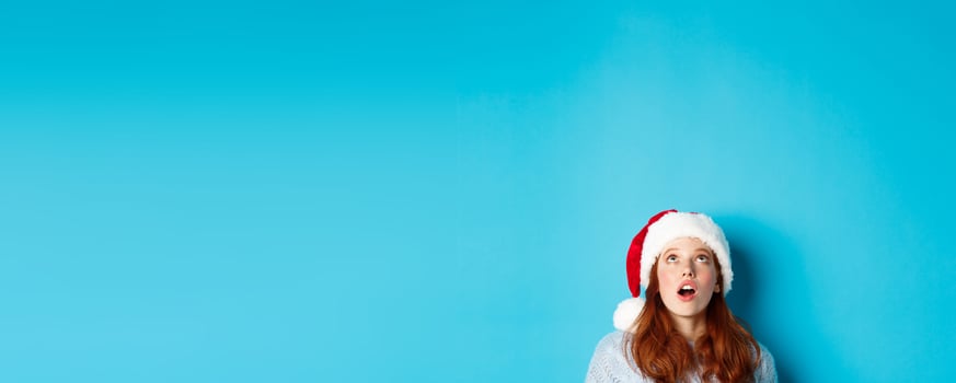 Winter holidays and Christmas eve concept. Head of pretty redhead girl in santa hat, appear from bottom and looking up at logo, seeing promo offer amazed, blue background photo