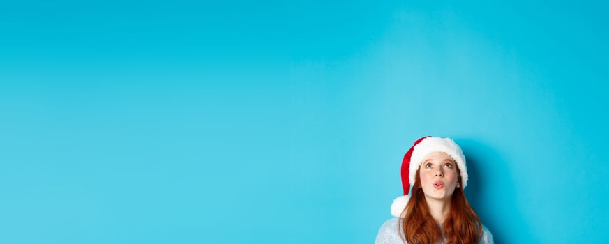 Winter holidays and Christmas eve concept. Head of pretty redhead girl in santa hat, appear from bottom and looking up at logo impressed, seeing promo offer, blue background photo