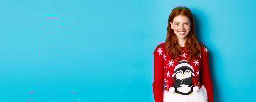 Winter holidays and Christmas Eve concept. Cute smiling teenage girl with red hair, wearing funny xmas sweater, standing against blue background photo