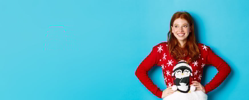 Winter holidays and Christmas Eve concept. Beautiful teenage redhead girl in xmas sweater looking left at logo, smiling pleased, holding hands on waist, blue background photo