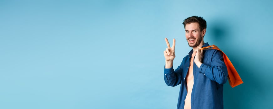 Cheerful guy holding orange shopping bag on shoulder, smiling and showing peace sign, standing on blue background photo