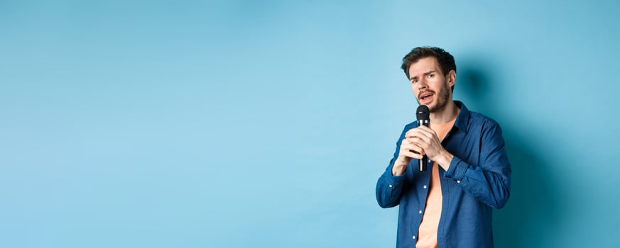 Young man singing song in microphone, playing karaoke, standing on blue background photo