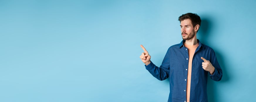 Image of young caucasian man looking and pointing aside, showing advertisement, standing on blue background photo