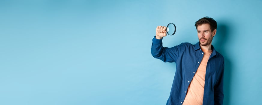 Image of young man searching for something, looking at empty space with magnifying glass, standing on blue background photo