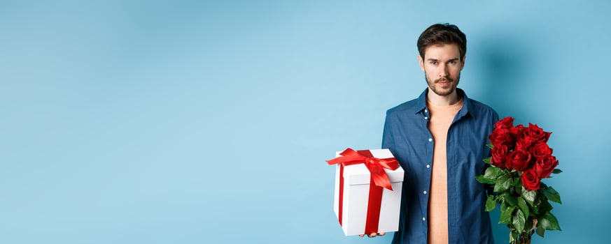 Love and Valentine day concept. Passionate man looking confident at camera, holding gift box and red roses for romantic date, standing over blue background photo