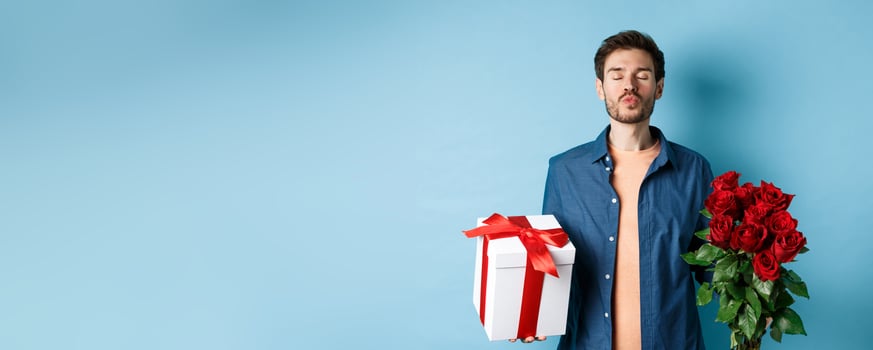 Love and Valentines day concept. Romantic man waiting for kiss, holding gift box and bouquet of red roses for lover on date, standing over blue background photo