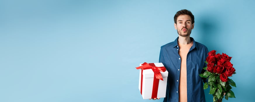 Love and Valentines day concept. Romantic boyfriend pucker lips for kiss, bring bouquet of red roses and gifts on date, standing over blue background photo