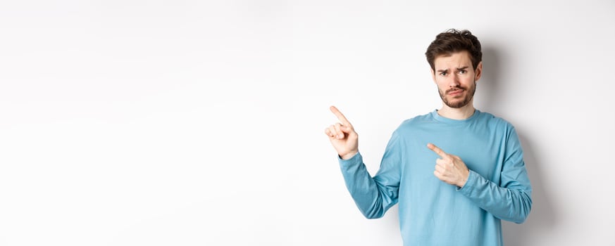 Skeptical young man with beard grimacing unsatisfied, frowning and pointing at upper left corner logo doubtful, standing upset over white background photo