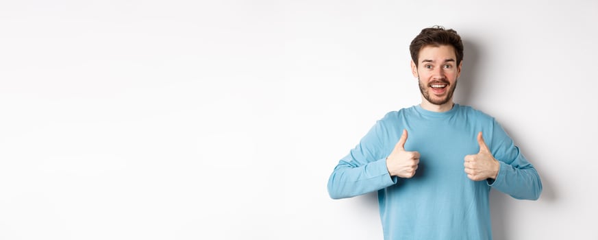 Excited smiling man showing thumbs up to praise excellent product, looking amazed and happy at camera, recommending good deal, standing on white background photo