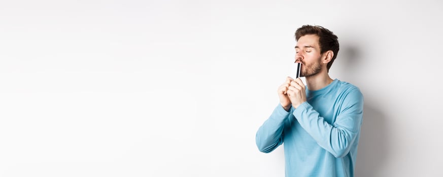 Young man kissing plastic credit card with satisfied face, standing over white background in blue casual shirt photo