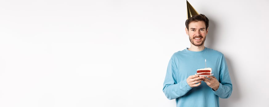 Celebration and holidays concept. Happy young man in party hat celebrating birthday, holding bday cake and smiling, standing over white background photo