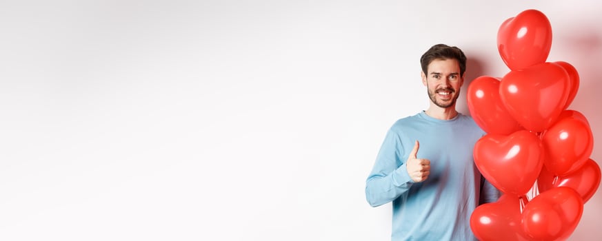 Smiling caucasian man standing with heart balloon, prepare surprise for lover on Valentines day, showing thumbs up and looking at camera, white background photo