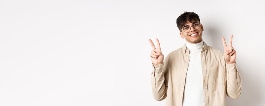 Handsome hipster guy in glasses, smiling with white teeth, showing peace or victory gesture, standing upbeat on white background photo
