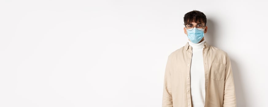 Young man in face mask from covid-19 looking cheerful at camera, standing on white background. Coronavirus, quarantine and social distancing concept photo