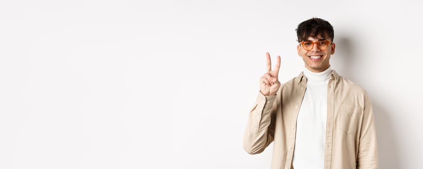 Handsome caucasian guy in glasses showing two fingers and smiling, standing on white background photo