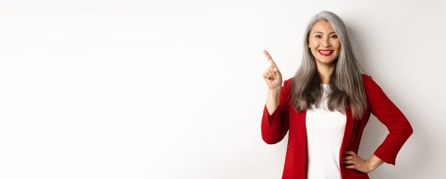 Happy senior office lady pointing finger at upper left corner, showing company logo and smiling at camera, standing over white background photo