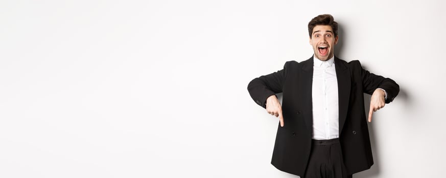 Portrait of super happy man showing christmas promo offer, pointing fingers down and smiling amazed, wearing formal party suit, standing over white background photo