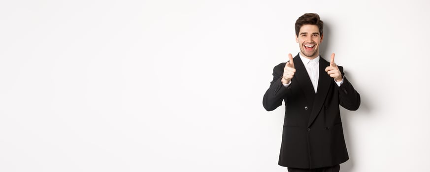 Concept of new year party, celebration and lifestyle. Handsome and successful guy in black suit, pointing fingers at camera and congratulating you, standing over white background photo