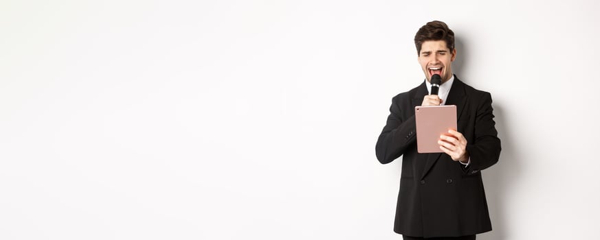 Image of handsome man in black suit, singing karaoke on digital tablet, holding microphone, standing over white background photo