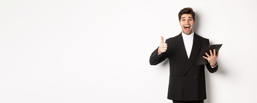 Portrait of confident businessman in black suit, holding clipboard with documents and showing thumb-up in approval, praise good job, standing against white background photo