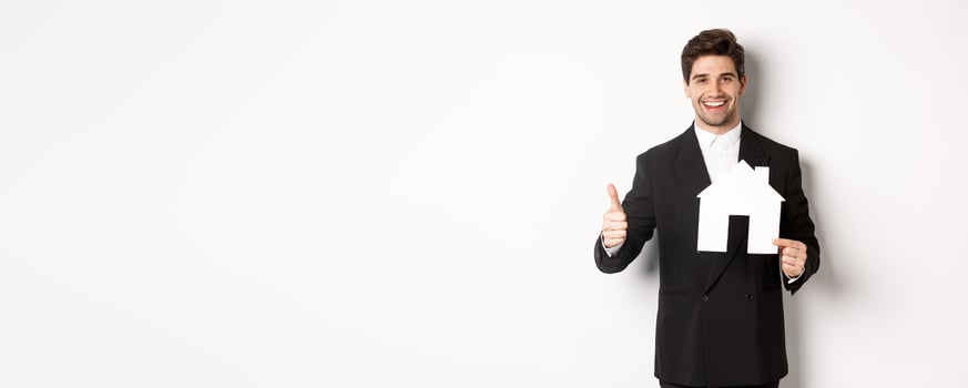 Portrait of confident real estate agent showing house maket and thumb-up, standing against white background photo