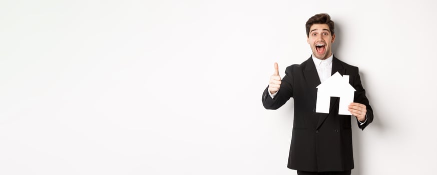 Portrait of excited handsome man looking for a home, showing house and thumb-up, recommending real estate agency, standing against white background photo