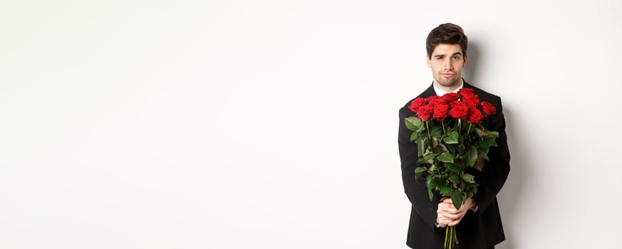 Image of handsome boyfriend in black suit, holding bouquet of red roses and smiling, being on a date, standing over white background photo