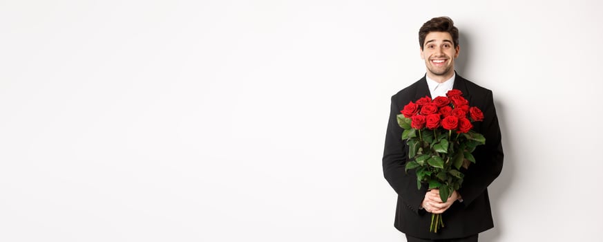 Image of handsome man in black suit, holding bouquet of roses and smiling, standing against white background photo