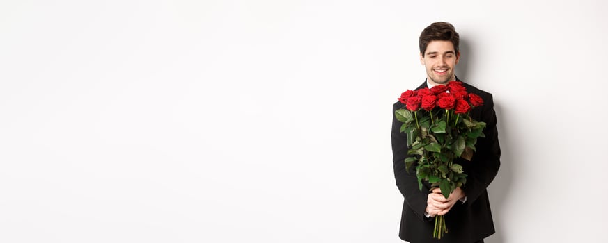 Image of handsome boyfriend in black suit, holding bouquet of red roses and smiling, being on a date, standing over white background photo