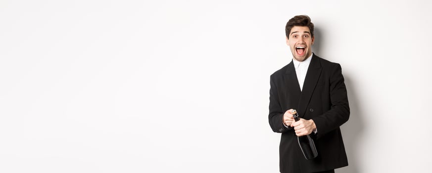 Image of attractive man in black suit having a party, celebrating new year and opening bottle of champagne, standing happy against white background photo
