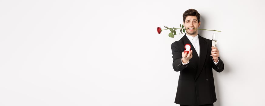 Romantic young man in suit making a proposal, holding rose in teeth and glass of champagne, showing engagement ring, asking to marry him, standing against white background photo