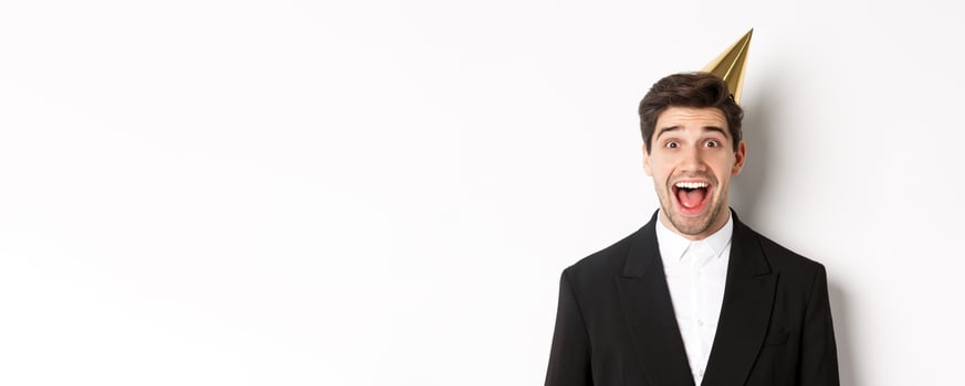 Close-up of excited handsome guy in trendy suit and party hat, looking amazed, celebrating winter holidays, standing against white background photo