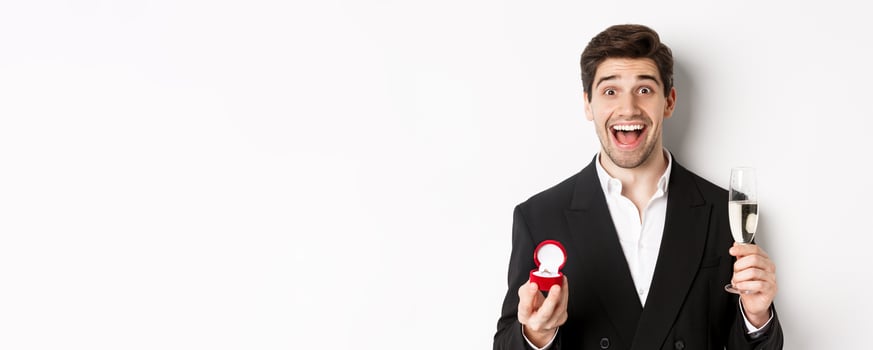 Close-up of handsome man in suit, making a proposal, giving engagement ring and raising glass of champagne, standing against white background photo