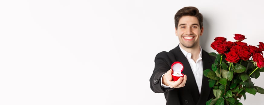 Close-up of attractive man in suit, holding bouquet of roses giving an engagement ring, proposing to girlfriend, standing against white background photo