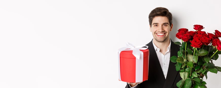 Close-up of handsome bearded man in suit, holding present and bouquet of red roses, smiling at camera, standing against white background photo