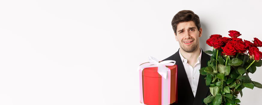 Close-up of skeptical man in suit, holding bouquet of red roses and a gift, standing reluctant against white background photo