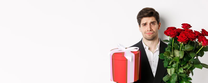 Close-up of sad man in suit, holding bouquet of red roses and a gift, standing upset against white background photo