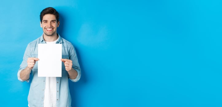 Young smiling guy in casual outfit, holding blank piece of paper with your advertisement, standing over blue background photo