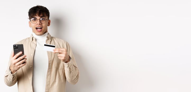Excited young man shopping online, holding mobile phone and plastic credit card, making purchase in internet, standing on white background photo