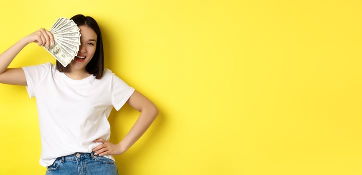 Cute asian woman hiding face behind money, peeking at camera satisfied, earn cash, standing over yellow background photo