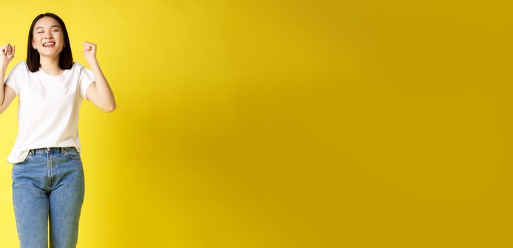 Full size shot of cheerful asian woman in jeans and white t-shirt jumping from happiness, winning prize and rejoicing, celebrating and shouting yes with joy, yellow background photo