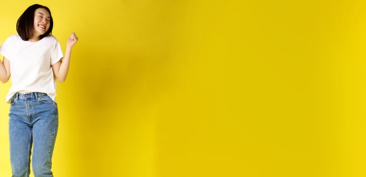 Full size shot of carefree asian woman jumping and dancing, having fun, posing in jeans and white t-shirt over yellow background photo