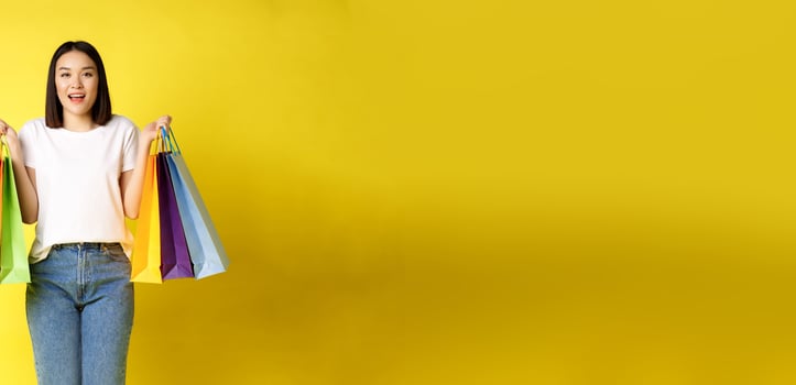 Full size portrait of beautiful asian girl going shopping, holding paper bags from stores and smiling, standing in jeans and white t-shirt over yellow background photo